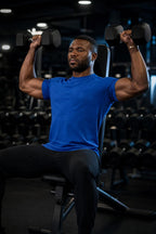 PrimeBuild Essential Training Tee in blue worn during a seated dumbbell shoulder press in a gym