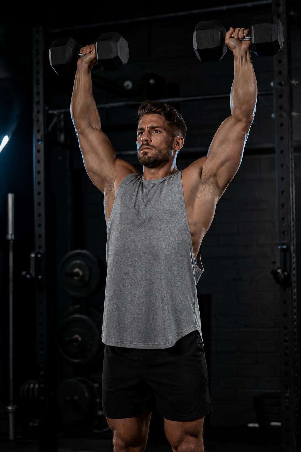 Man wearing the PrimeBuild Men's Stringer Gym Tank in gray, crew neck, pressing two dumbbells overhead in a gym