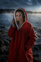 Woman wearing the red PrimeBuild Swim Parka with its sherpa-lined hood up on a rocky shore by misty water