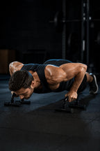 PrimeBuild Push-Up Bars in use: man in a black tank top doing a push-up on a pair of black push-up bars on a dark gym floor