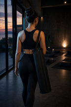 PrimeBuild Yoga Mat Carry Bag in black carried over the shoulder by a woman walking through a dimly lit yoga studio