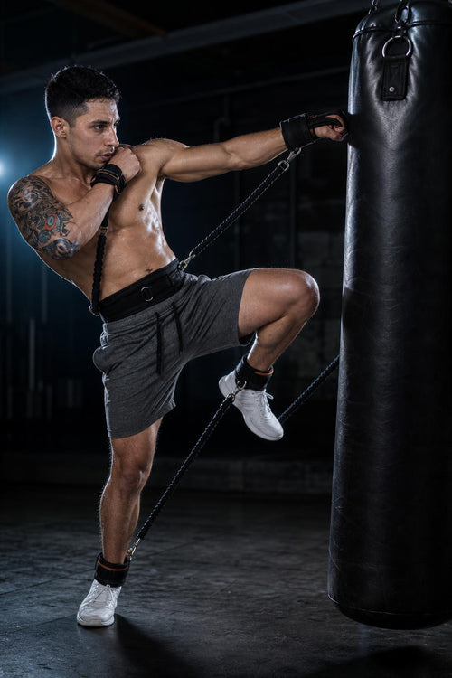 Athlete throwing a knee strike at a heavy bag wearing the PrimeBuild Combat Resistance Training Set belt, bands and cuffs