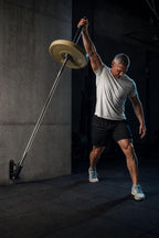 PrimeBuild Wall-Mounted Landmine Attachment on a concrete wall holding a loaded barbell during a one-arm press