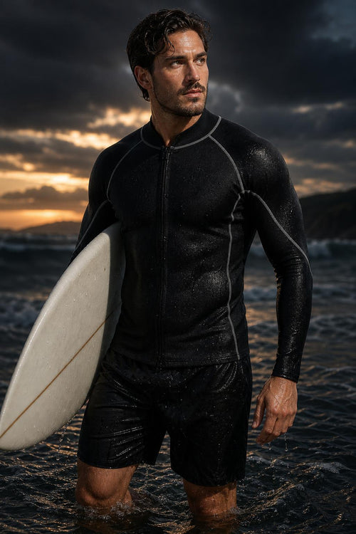 Man wearing the PrimeBuild 2mm neoprene wetsuit top, standing in the ocean at sunset and holding a white surfboard