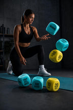 PrimeBuild Exercise Dice in use: a woman kneeling on a teal mat tosses blue and yellow foam dice in a dark workout room