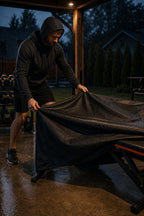 Man pulling the black PrimeBuild Weight Bench Cover over a red-trimmed bench on a covered patio in the rain at dusk