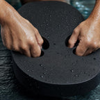 Close-up of wet hands gripping the hand holes of a black PrimeBuild swim kickboard, water drops on the foam