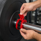 Hands fitting a red PrimeBuild Olympic Barbell Collar onto a barbell sleeve next to a black weight plate, lever raised